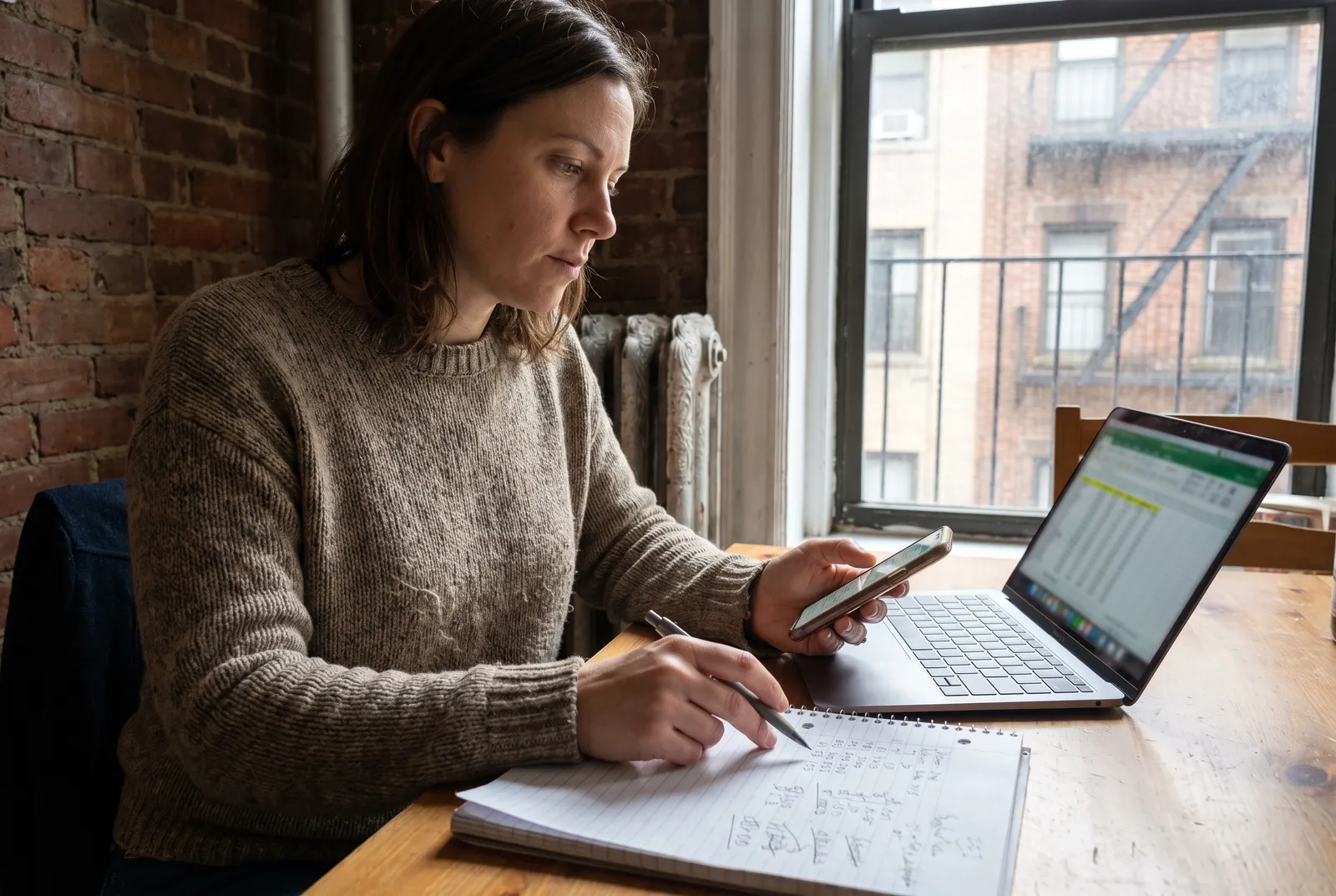 Woman comparing dog care prices on phone and notebook in New York City apartment