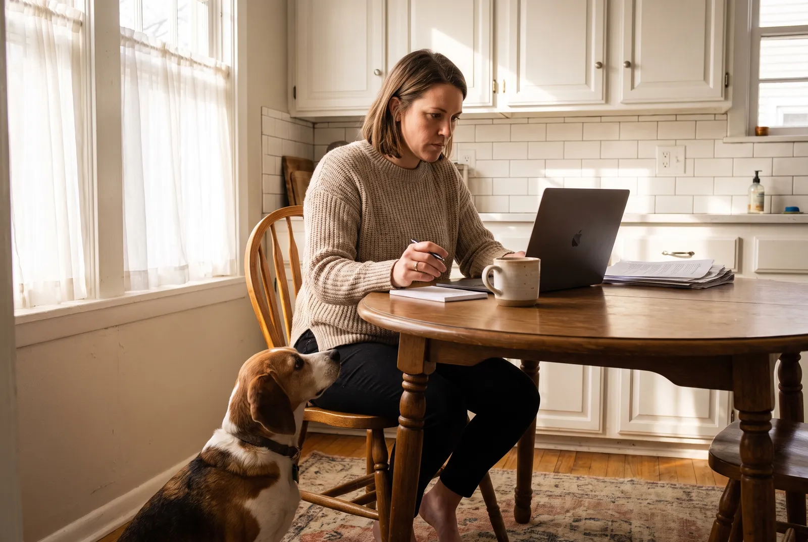 A woman at a bright kitchen table reviews options on a laptop while her beagle mix sits on the floor beside her.
